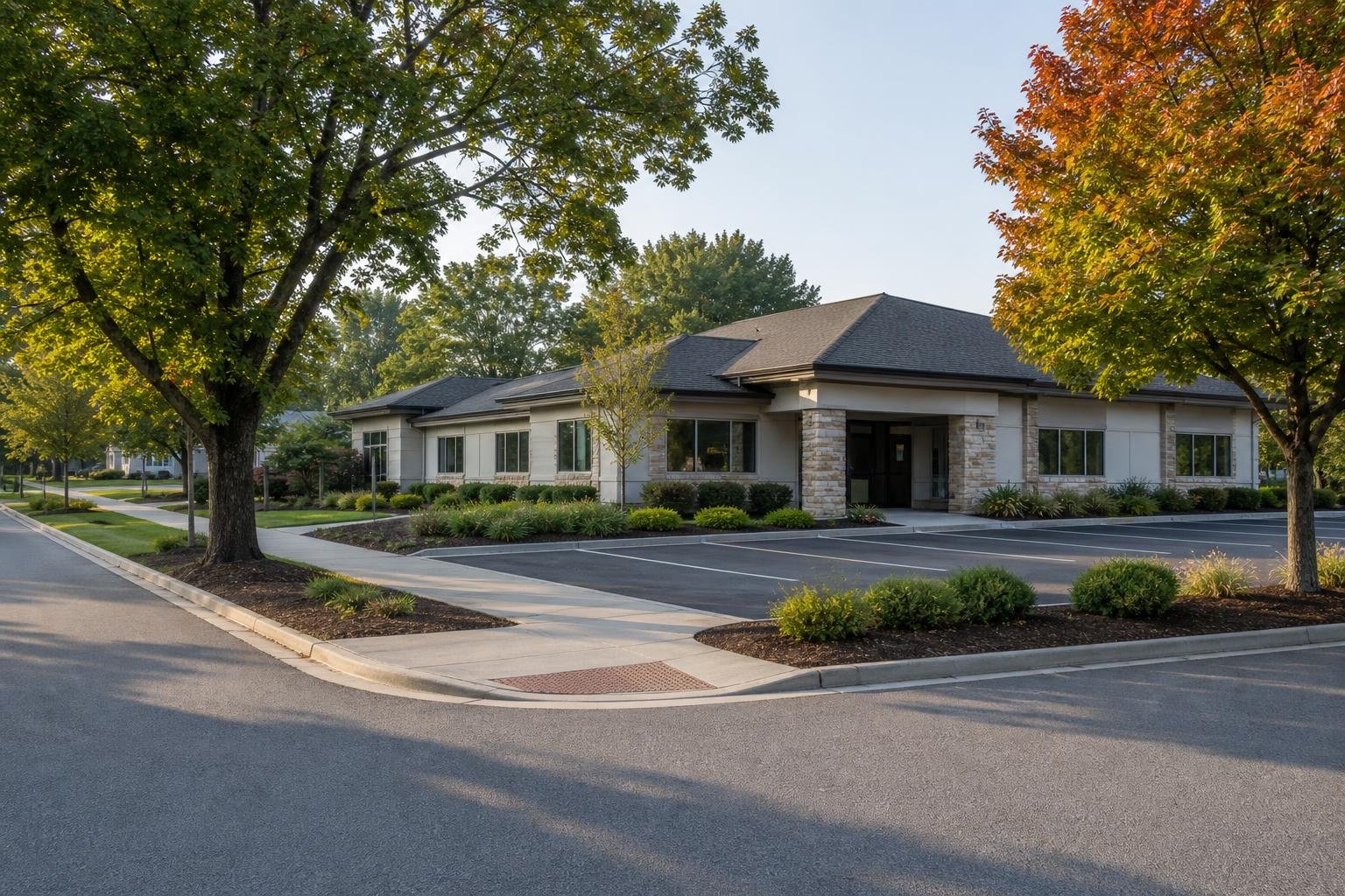 A low, single-story commercial building sits beside an empty parking lot and street, surrounded by trees with green and orange leaves.